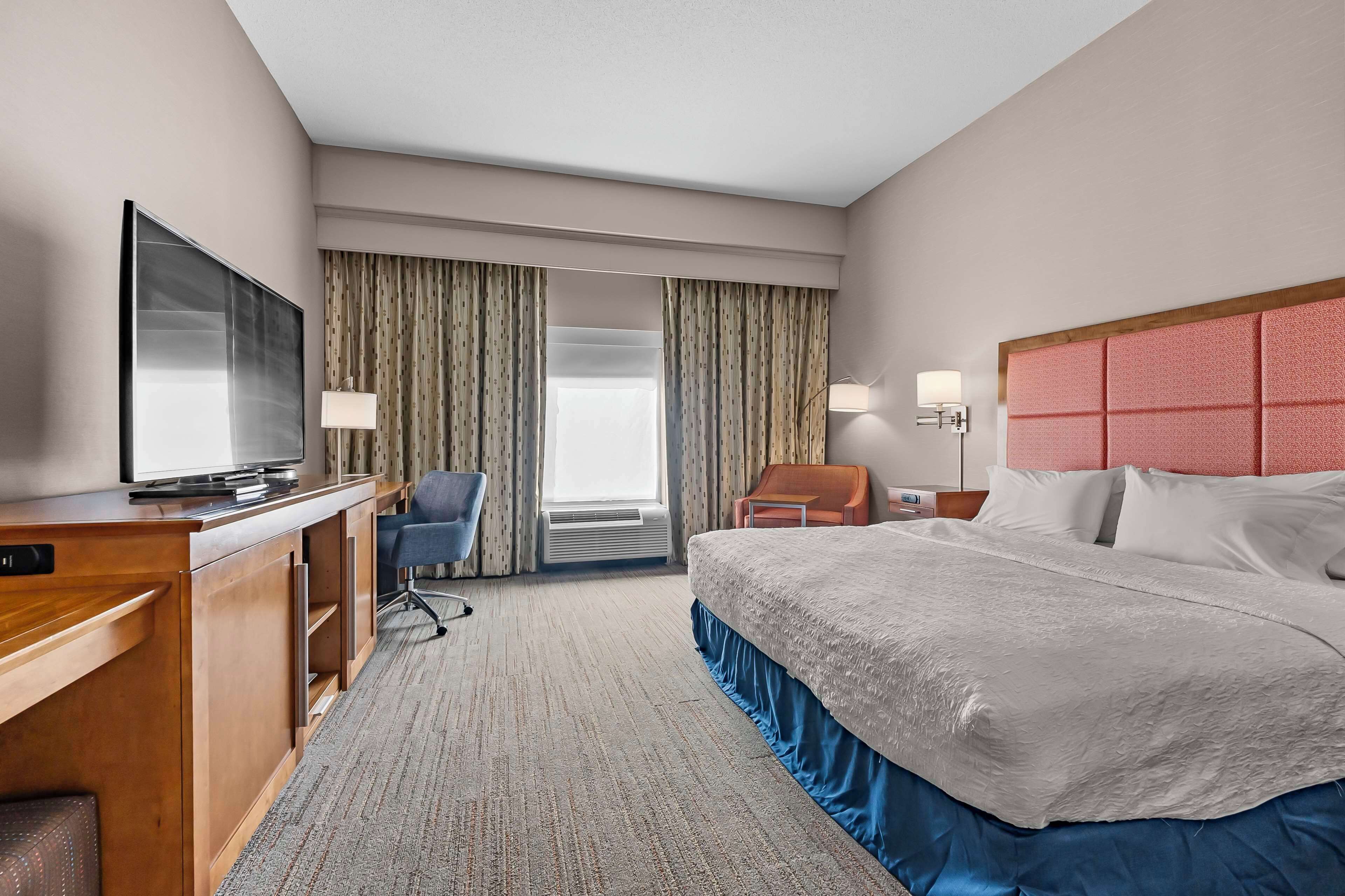 Une chambre d’hôtel comprenant un lit, un bureau avec une télévision, une chaise, une fenêtre avec des rideaux et une table de chevet avec une lampe.