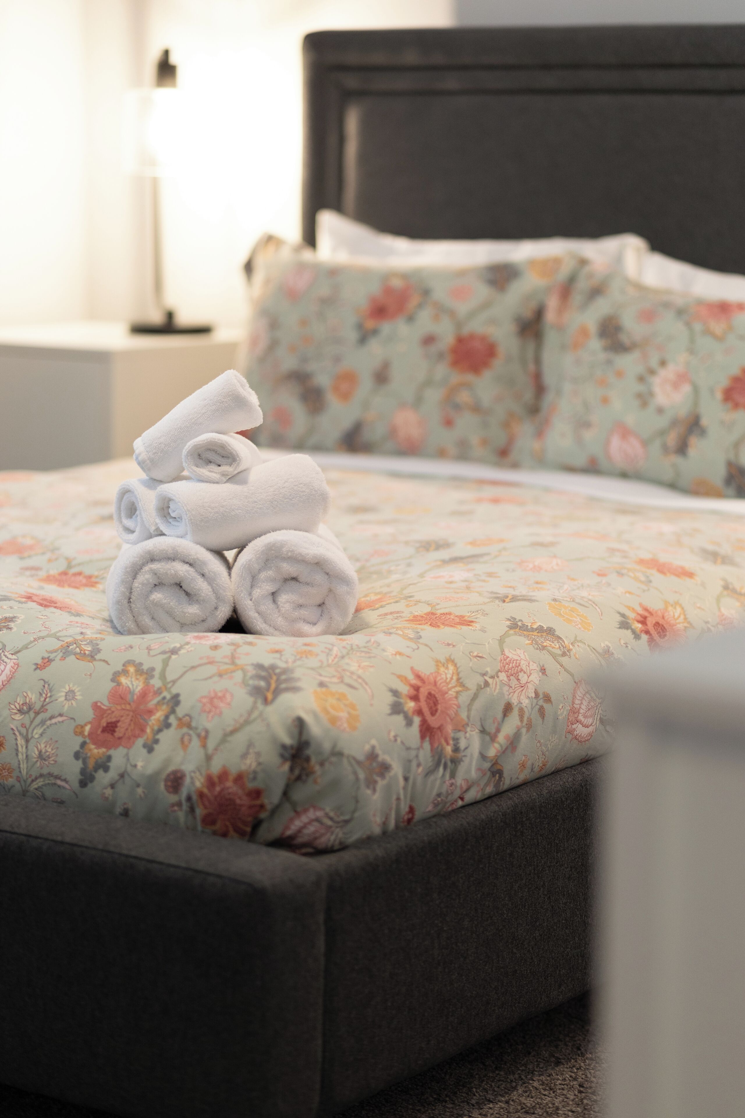 Una cama bien hecha con ropa de cama floral, una pila de toallas enrolladas y un cabecero oscuro.