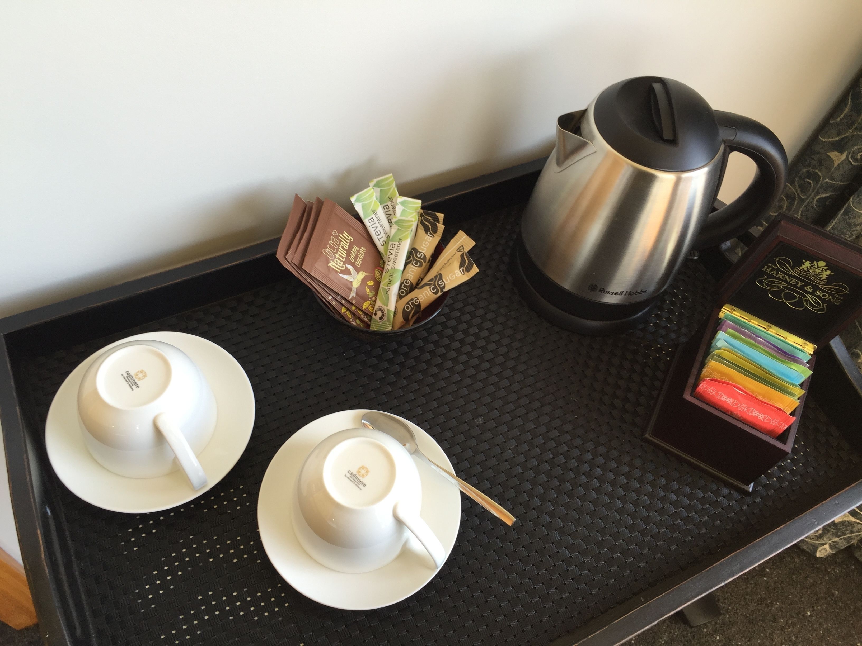 A tray with two white cups and saucers, a stack of organic sugar packets, a black coffee pot, and a box of assorted chocolates.