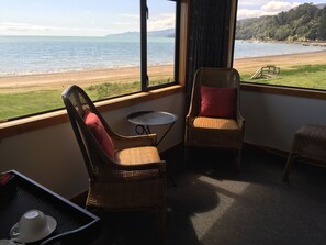 A room with wicker chairs, a small round table, and a view of a beach and mountains. - Te Puru Beach Lodge (Te Puru)