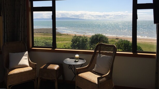 Ein Zimmer mit Korbstühlen und einem kleinen Tisch, ein großes Fenster mit Blick auf Strand und Berge.