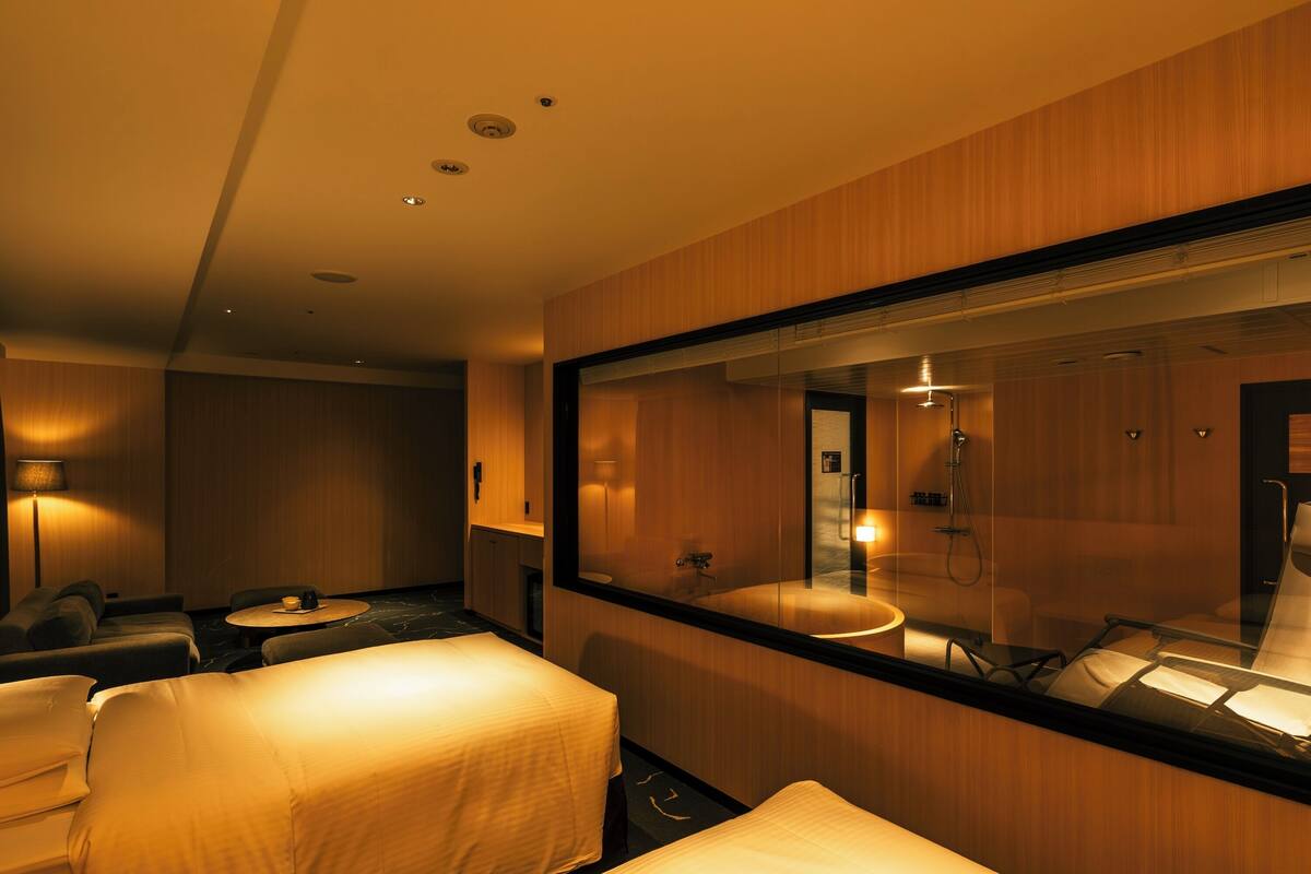 Habitación de hotel con dos camas, una zona de estar con una mesa y un espejo grande que refleja el baño.