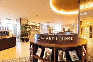 A modern lounge area with a 'Share Lounge' sign, wooden shelves with books, and a large potted plant.