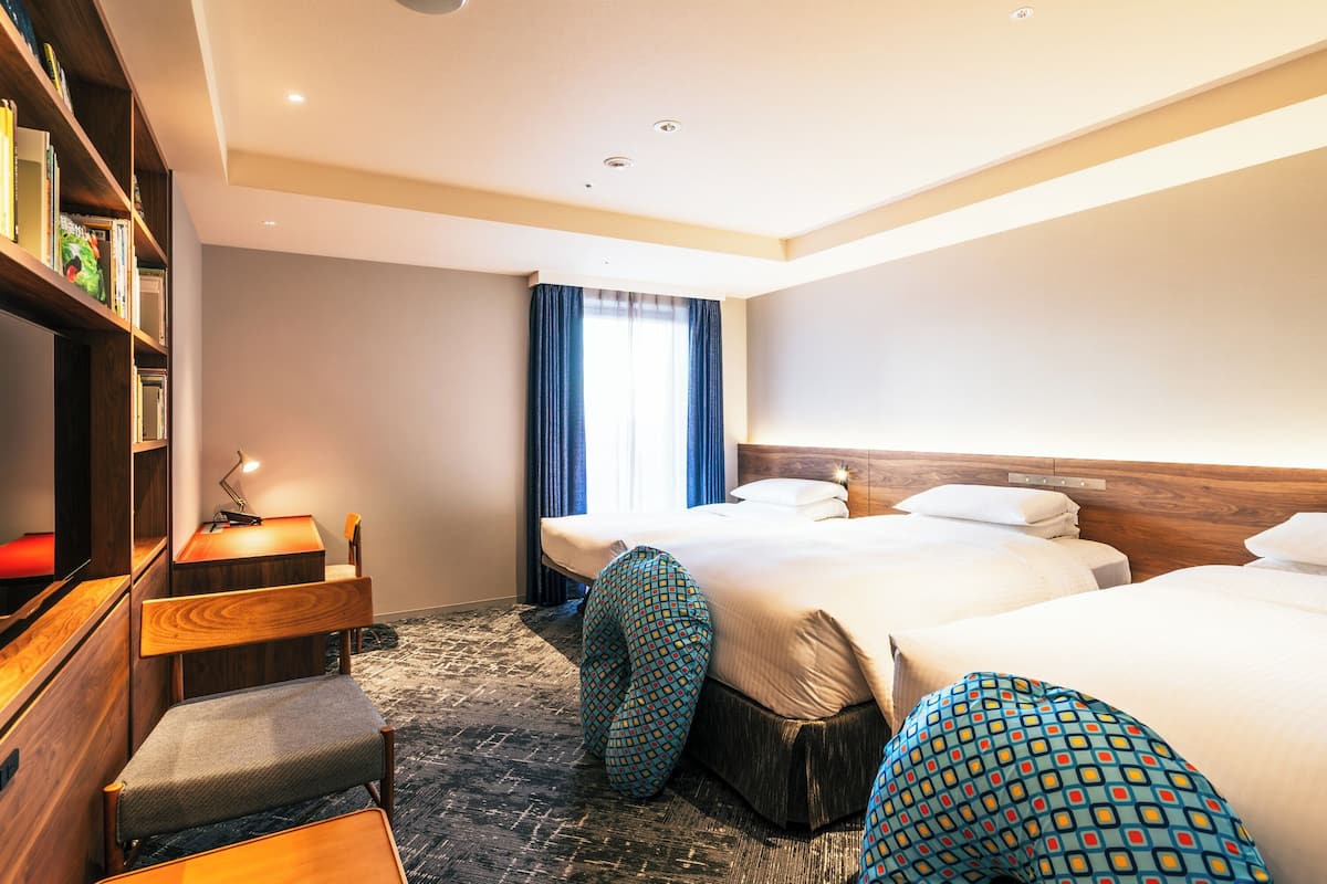Habitación de hotel con dos camas, un escritorio con lámpara, una estantería y un ventanal con cortinas.