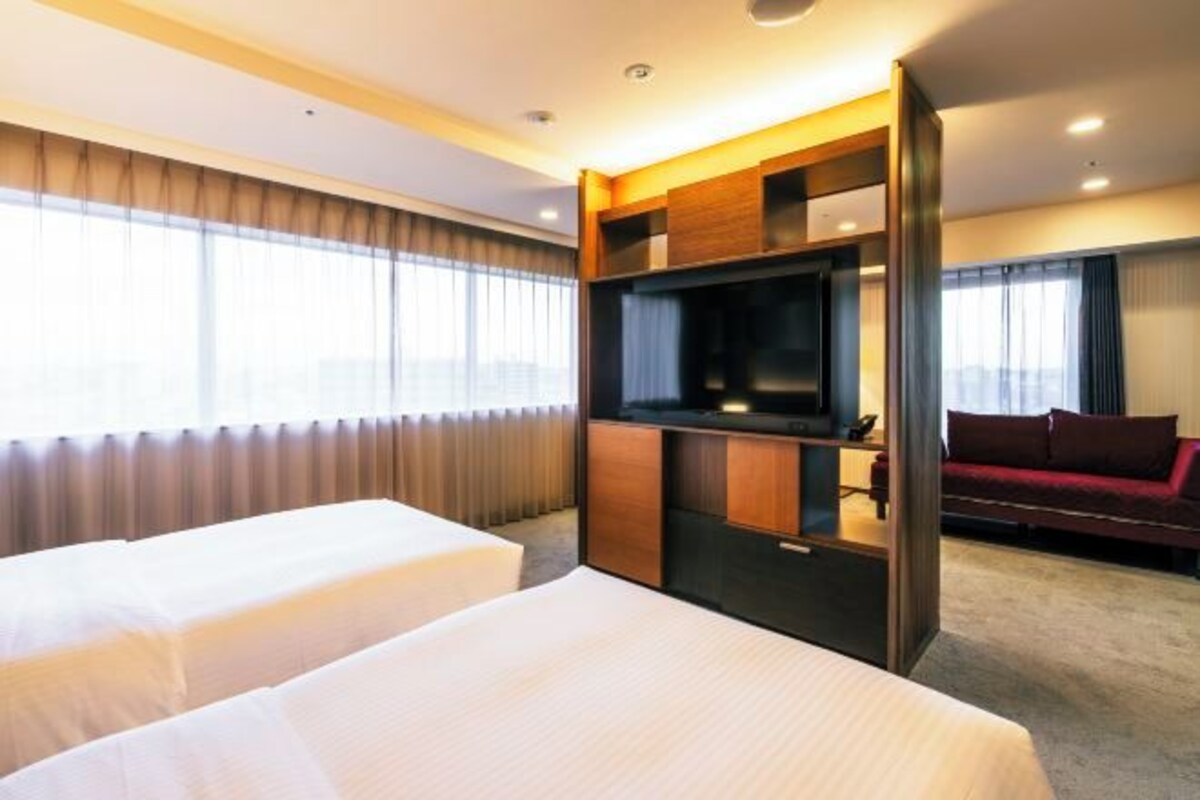 Habitación de hotel con dos camas, un televisor, un sofá rojo y un ventanal con cortinas.