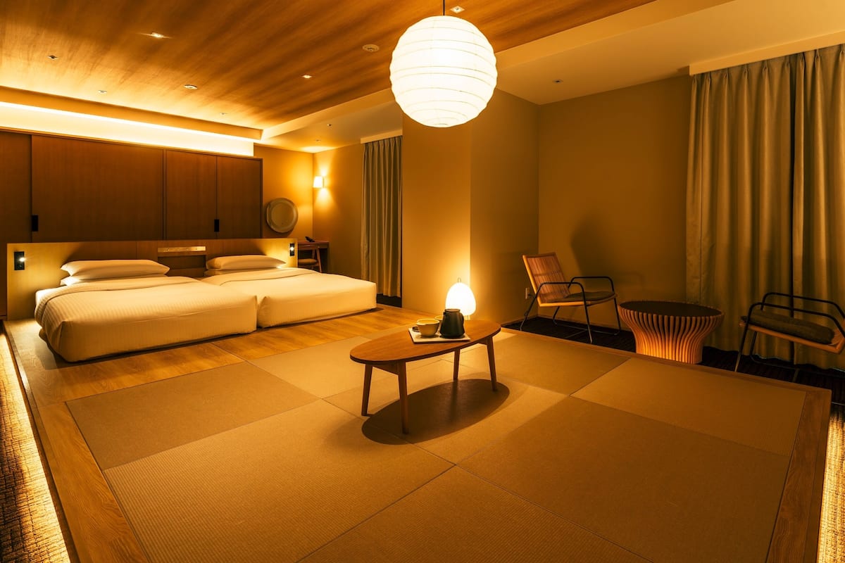 Una habitación de hotel moderna con una cama grande, un espacio de descanso con una mesa y sillas, y vistas de la ciudad a través de la ventana.