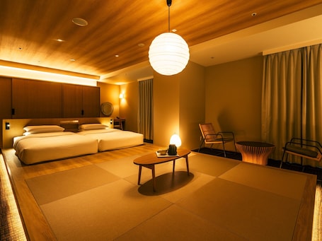 A modern hotel room with a large bed, a sitting area with a table and chairs, and a view of the city through the window.