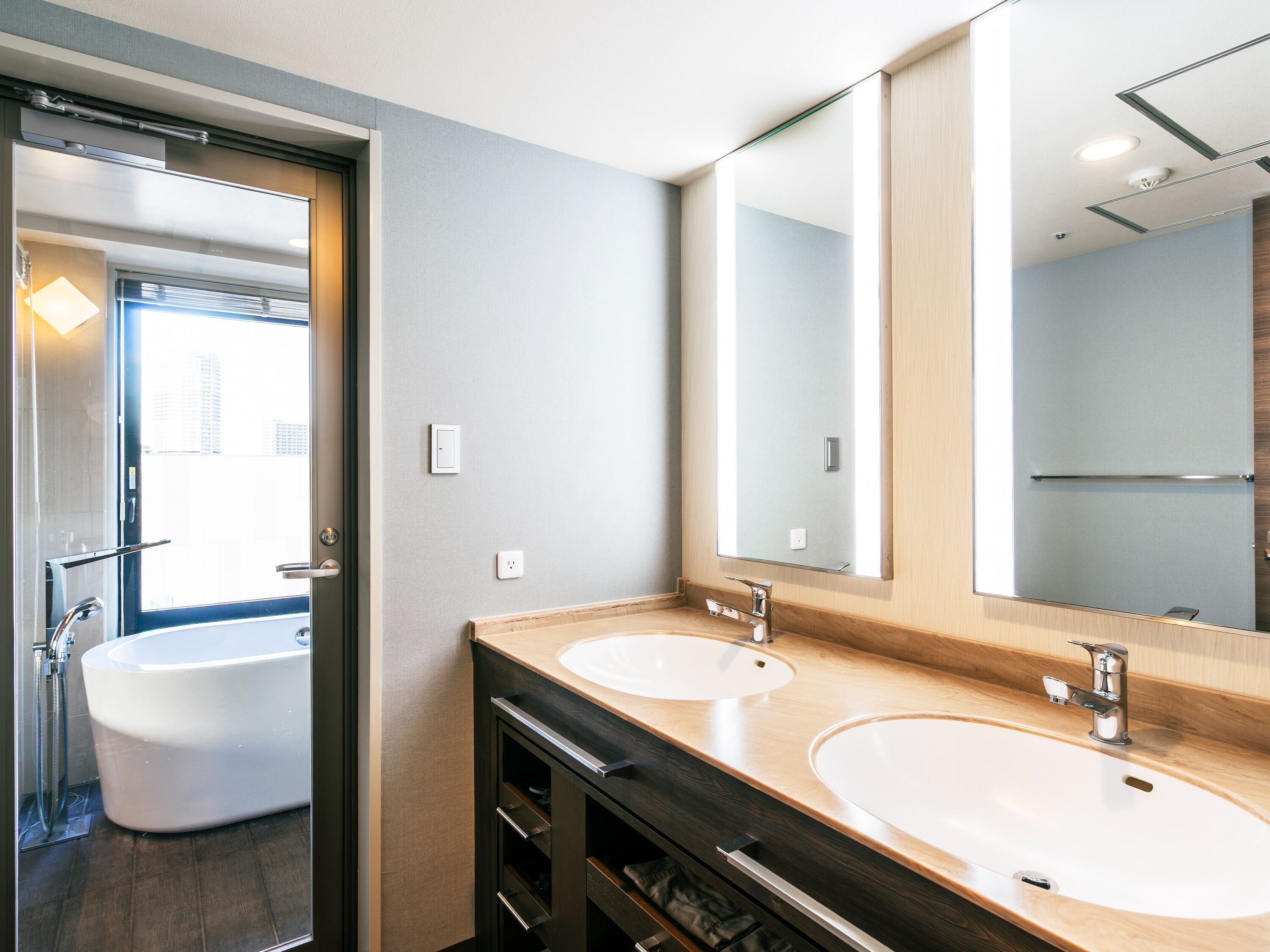 A modern bathroom with two sinks, a large mirror, and a bathtub visible through an open door.