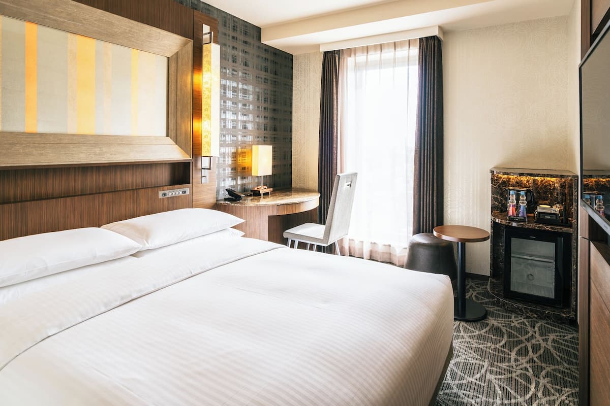 Habitación de hotel con una cama grande, un escritorio con silla, una mesita pequeña, un minibar y un ventanal con cortinas.