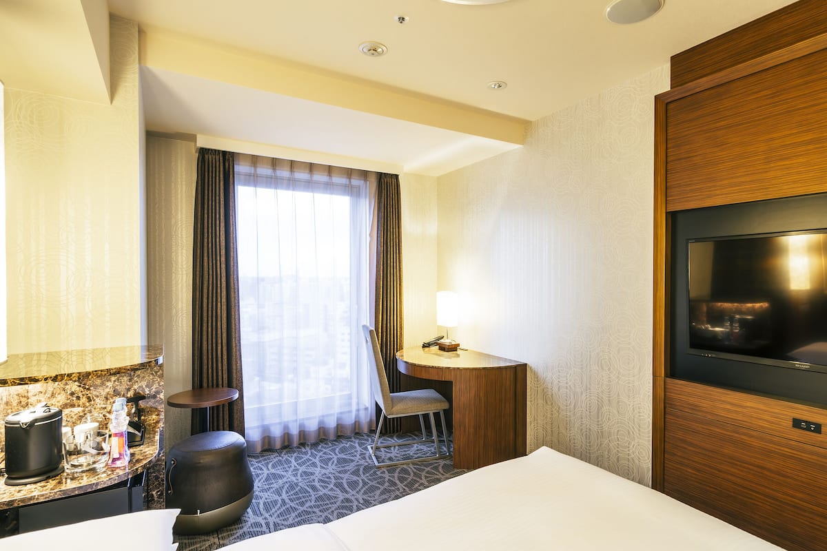 Una habitación de hotel con cama, escritorio con silla, televisor y ventana con cortinas.