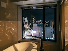 A modern bathroom with a bathtub, a large window with a city view, and a toilet.