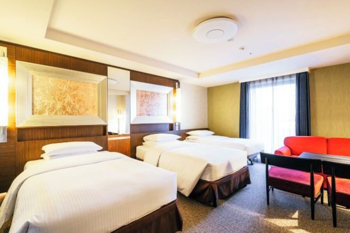 Habitación de hotel con dos camas, un sofá rojo, un escritorio y un ventanal grande.