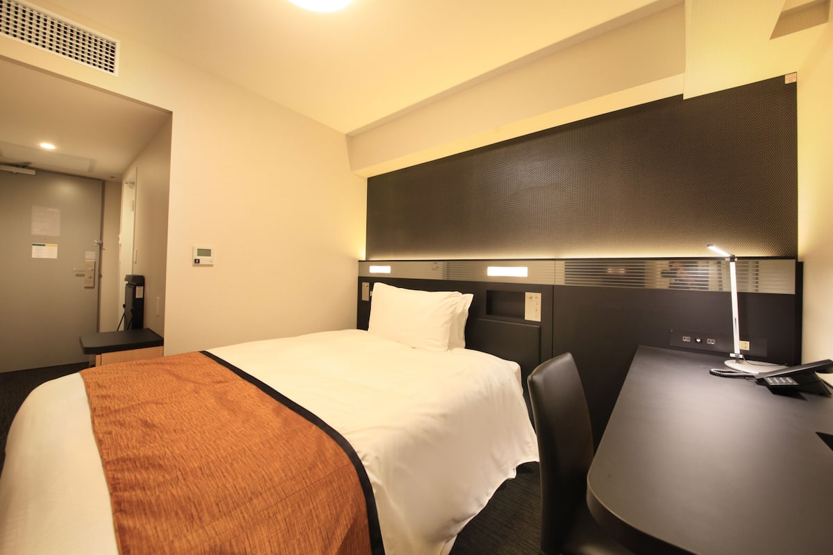 A hotel room with a bed, a desk, a chair, a desk lamp, and a telephone.