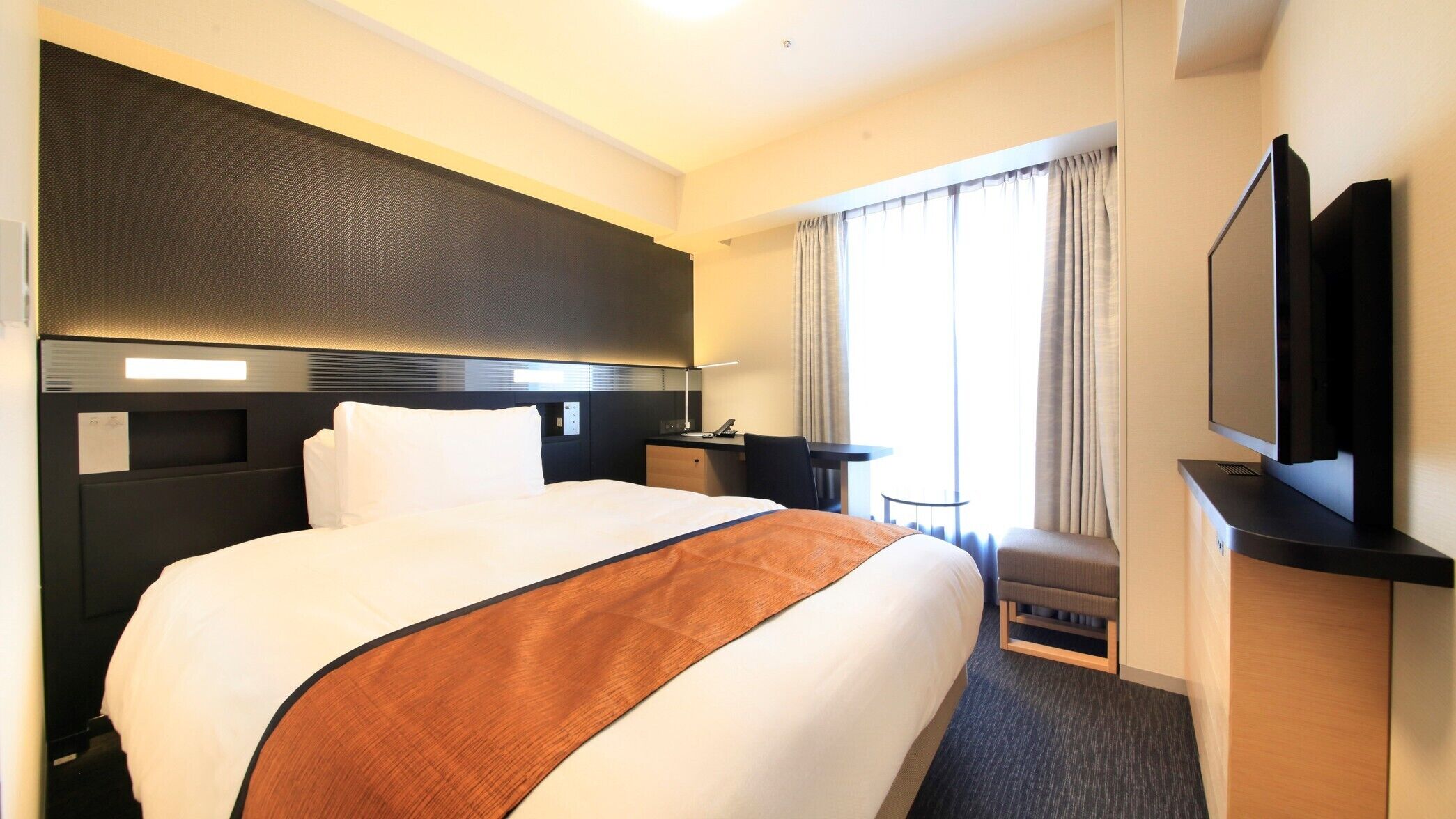 A hotel room with a large bed, a desk, a chair, a television, and a window with curtains.