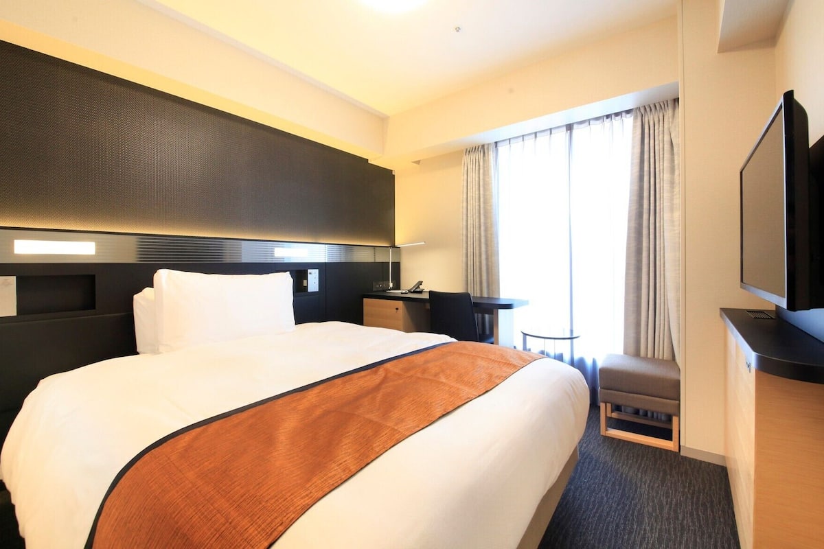 Une chambre d’hôtel avec un grand lit, un bureau, une chaise, une télévision et une fenêtre avec des rideaux.