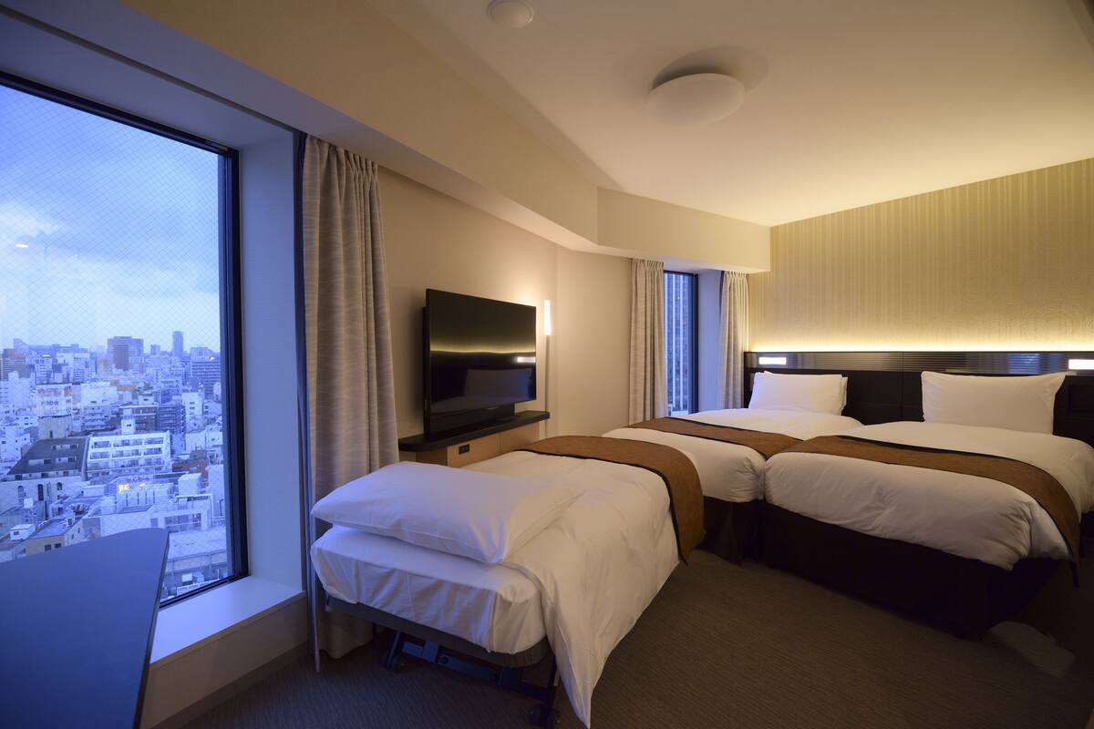 A hotel room with a large window, a flat-screen TV, two beds, and a city view.