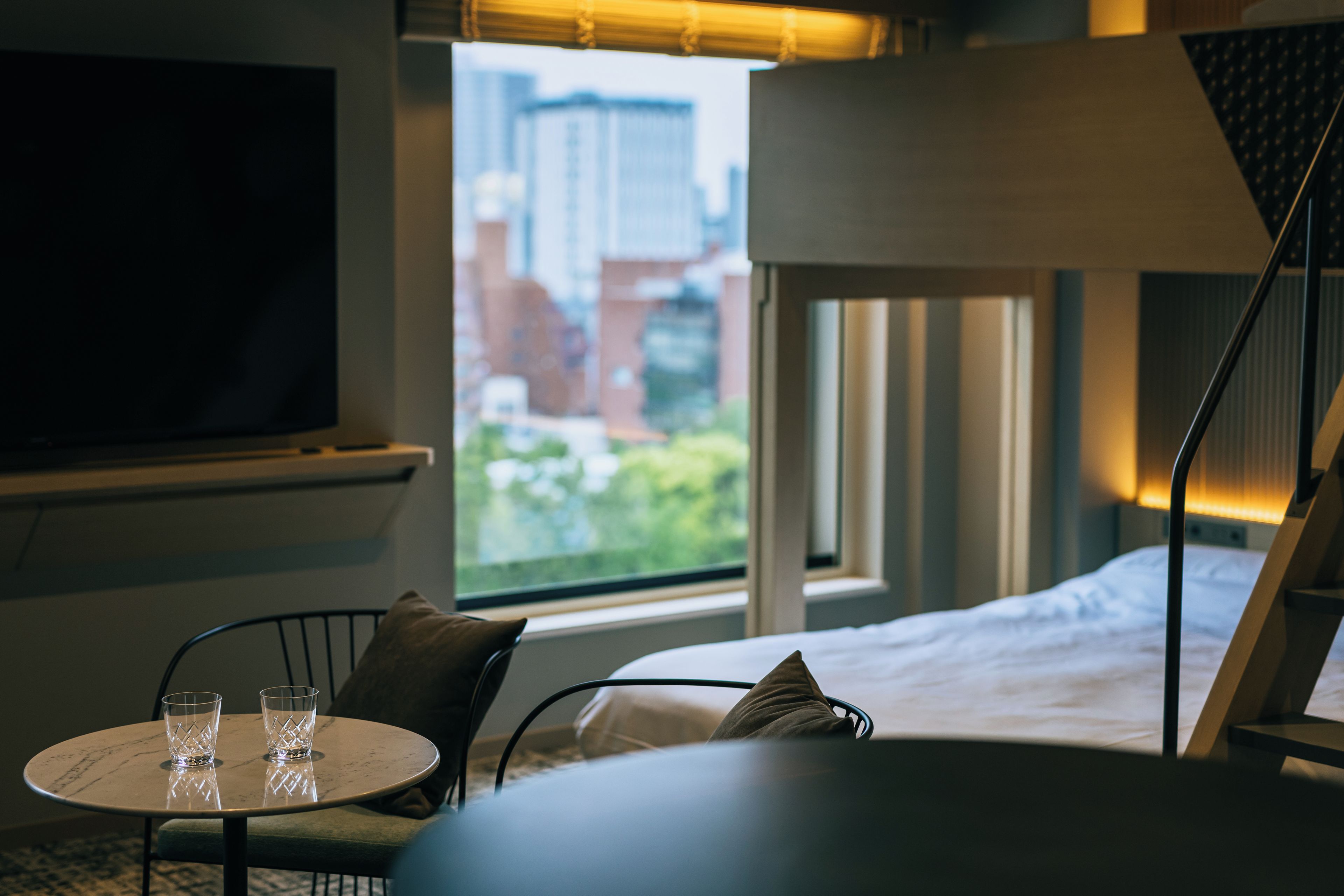 Quarto de hotel com cama, televisão, mesa com copos e janela vista para edifícios.
