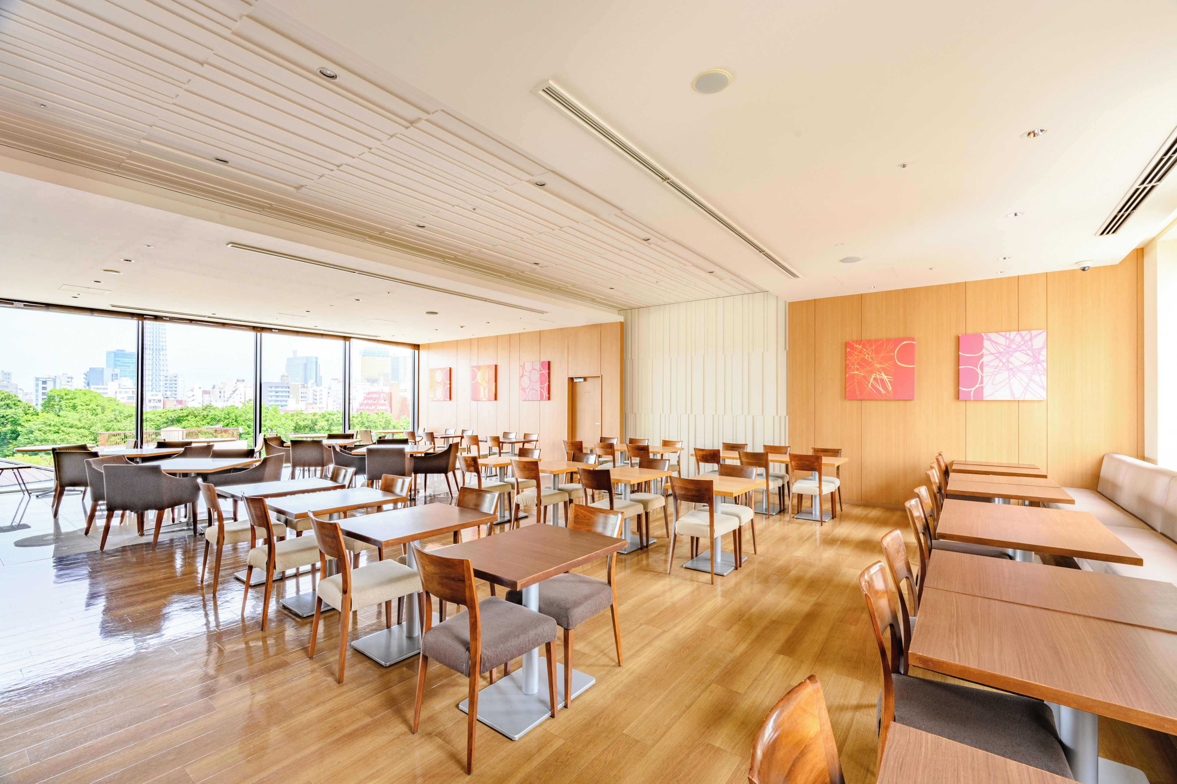 A spacious dining area with multiple tables and chairs, large windows offering a view of the city, and a high ceiling with linear lighting fixtures.