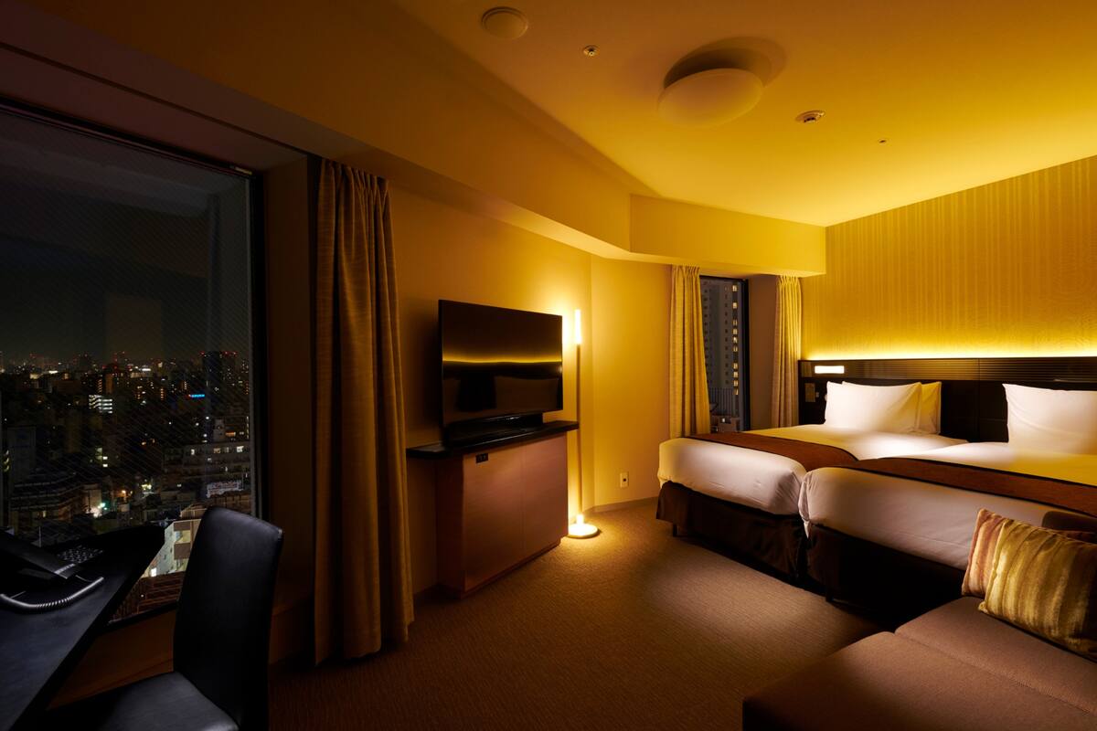 A hotel room with a large window, a desk, a bed, and a sofa.