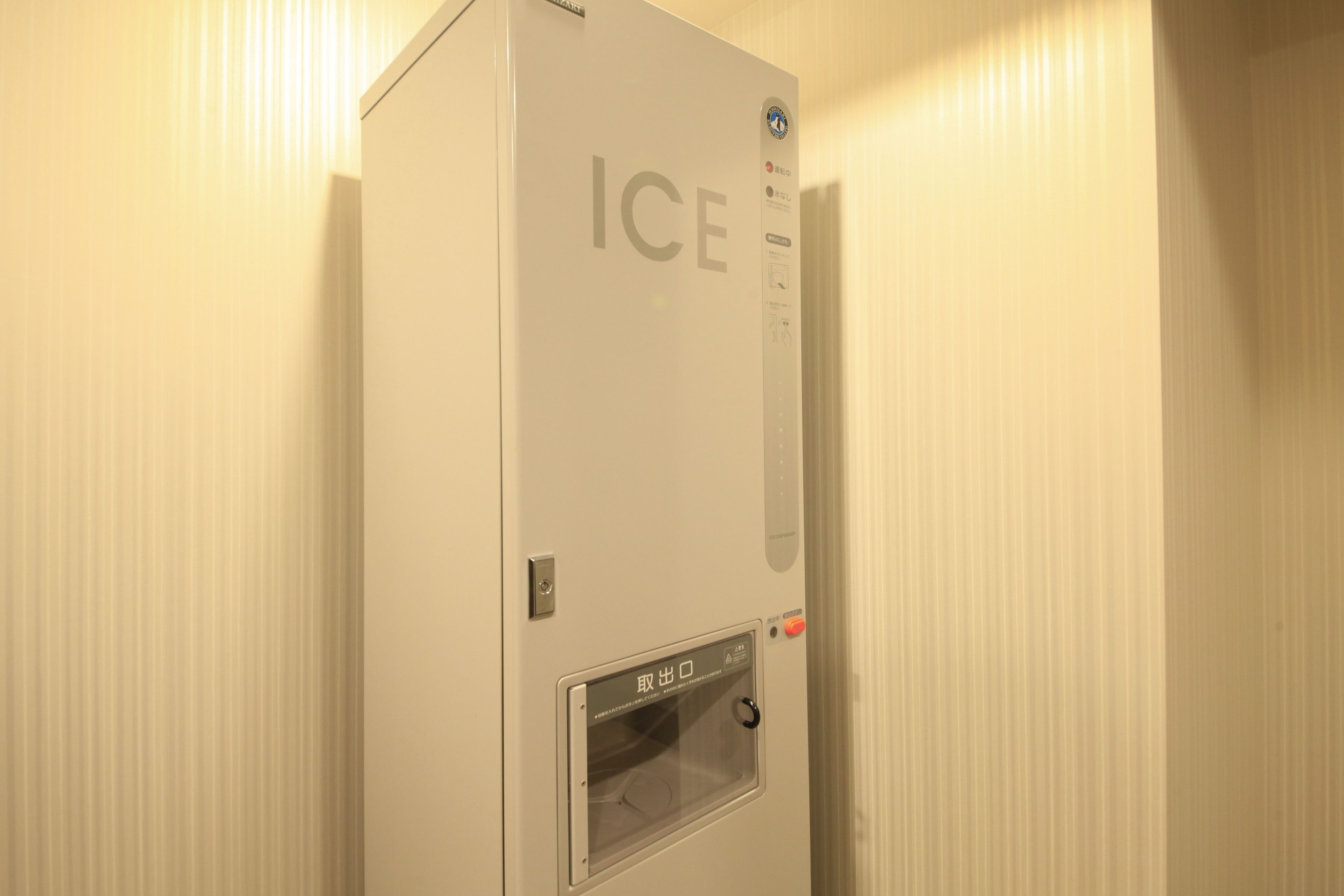 Una máquina de hielo blanca con la palabra “ICE” en la parte frontal, un panel de control con botones y una pequeña ventana.