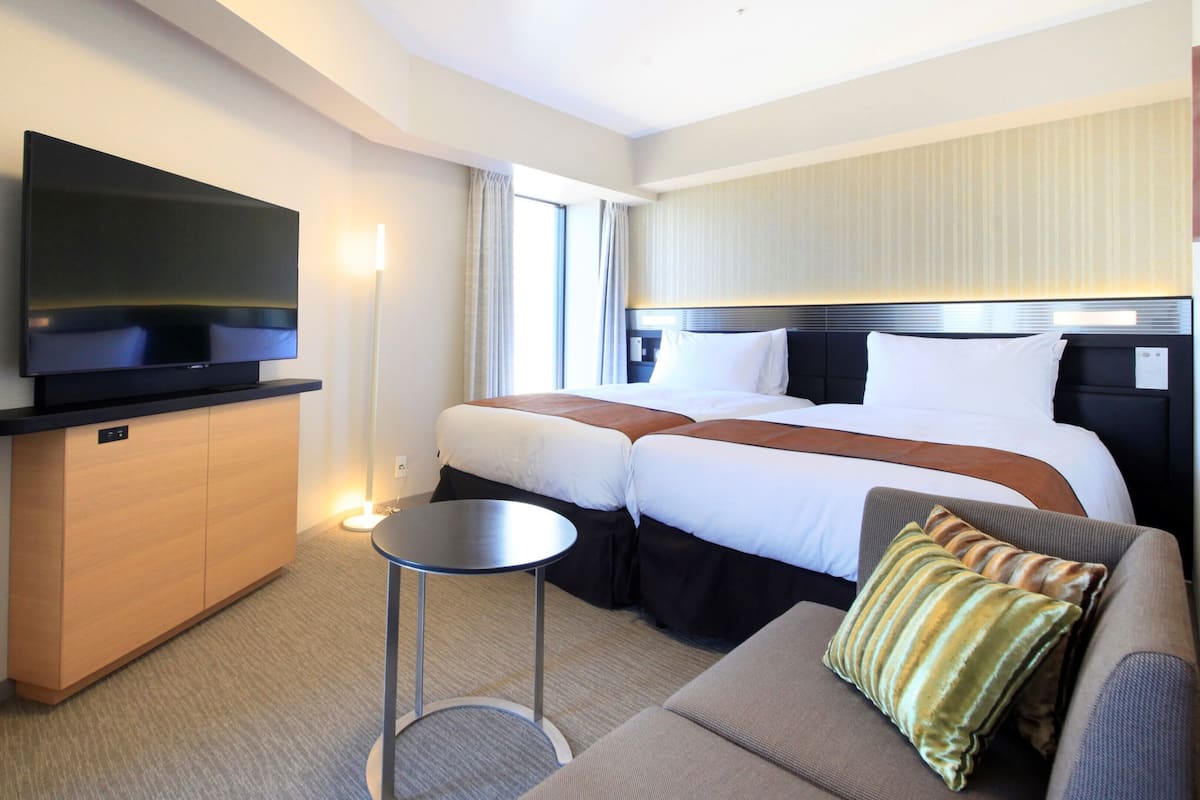 A hotel room with a flat-screen TV, two beds, a sofa, a small table, and a desk with a flat-screen TV.