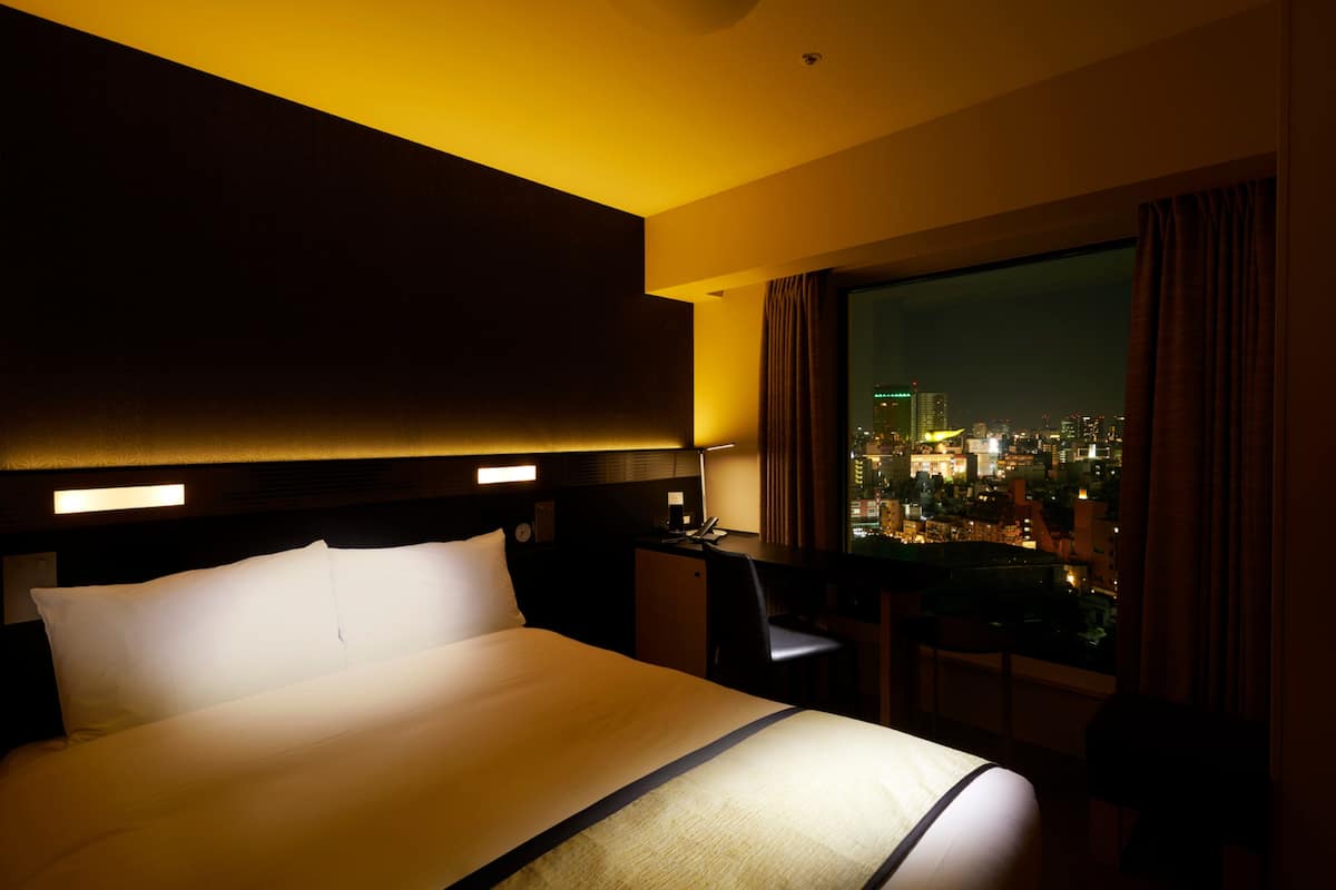 A hotel room with a large bed, a desk, and a view of a cityscape at night.