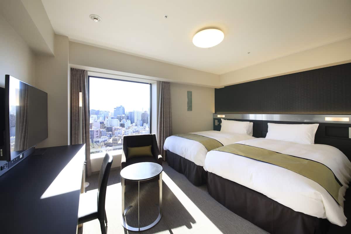 A hotel room with two beds, a desk, a chair, a TV, and a large window with a city view.