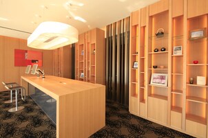 Egy modern szállodai előcsarnok, amelyben található egy recepciós pult, egy ülősarok és beépített, különböző tárgyakkal teli fa polcrendszer.