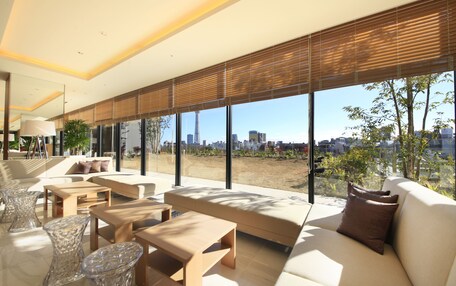A modern living room with a sofa, coffee table, and chairs, large windows with blinds, and a city view.