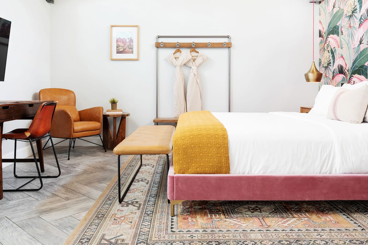 A hotel room with a bed, a desk, a chair, a small table with a plant, and a wall-mounted coat rack.
