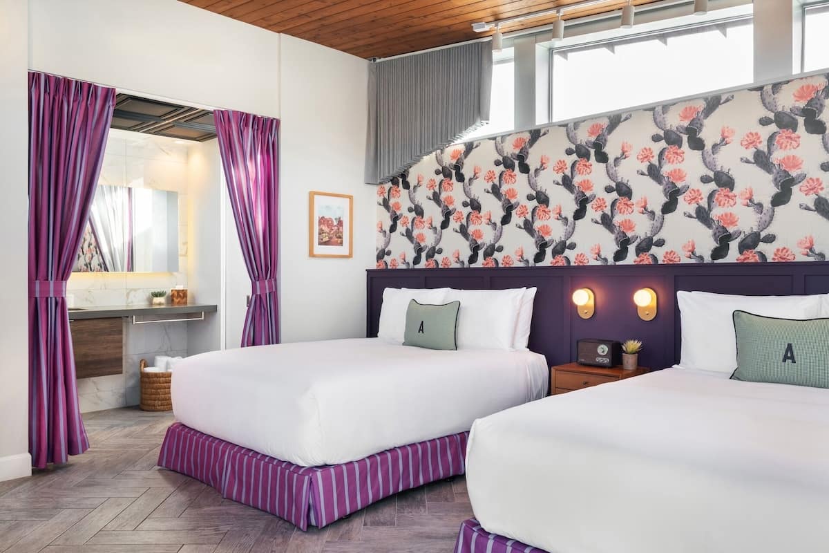 A hotel room with two beds, a bathroom with a mirror, and a floral wallpaper.