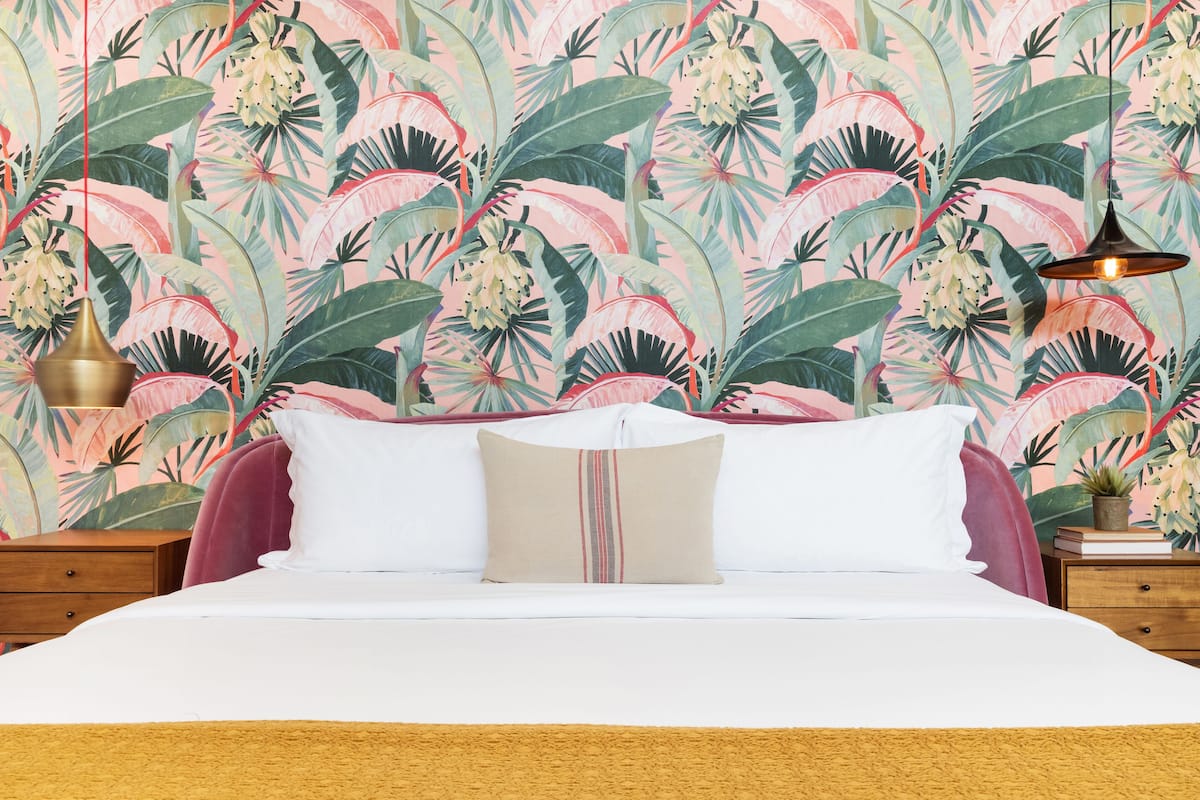 A bed with white bedding, a beige pillow, and a pink headboard. There is a nightstand with a lamp and a small plant. The wall features a tropical leaf pattern.