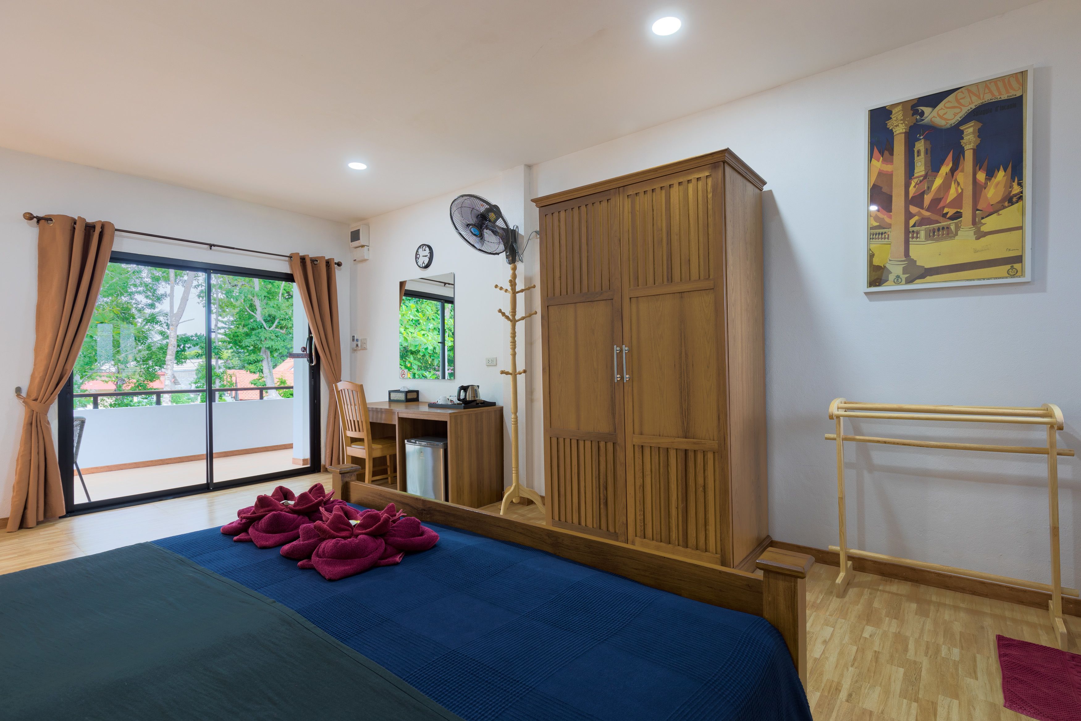 A room with a bed, a wooden wardrobe, a desk, a chair, a fan, and a window with curtains.