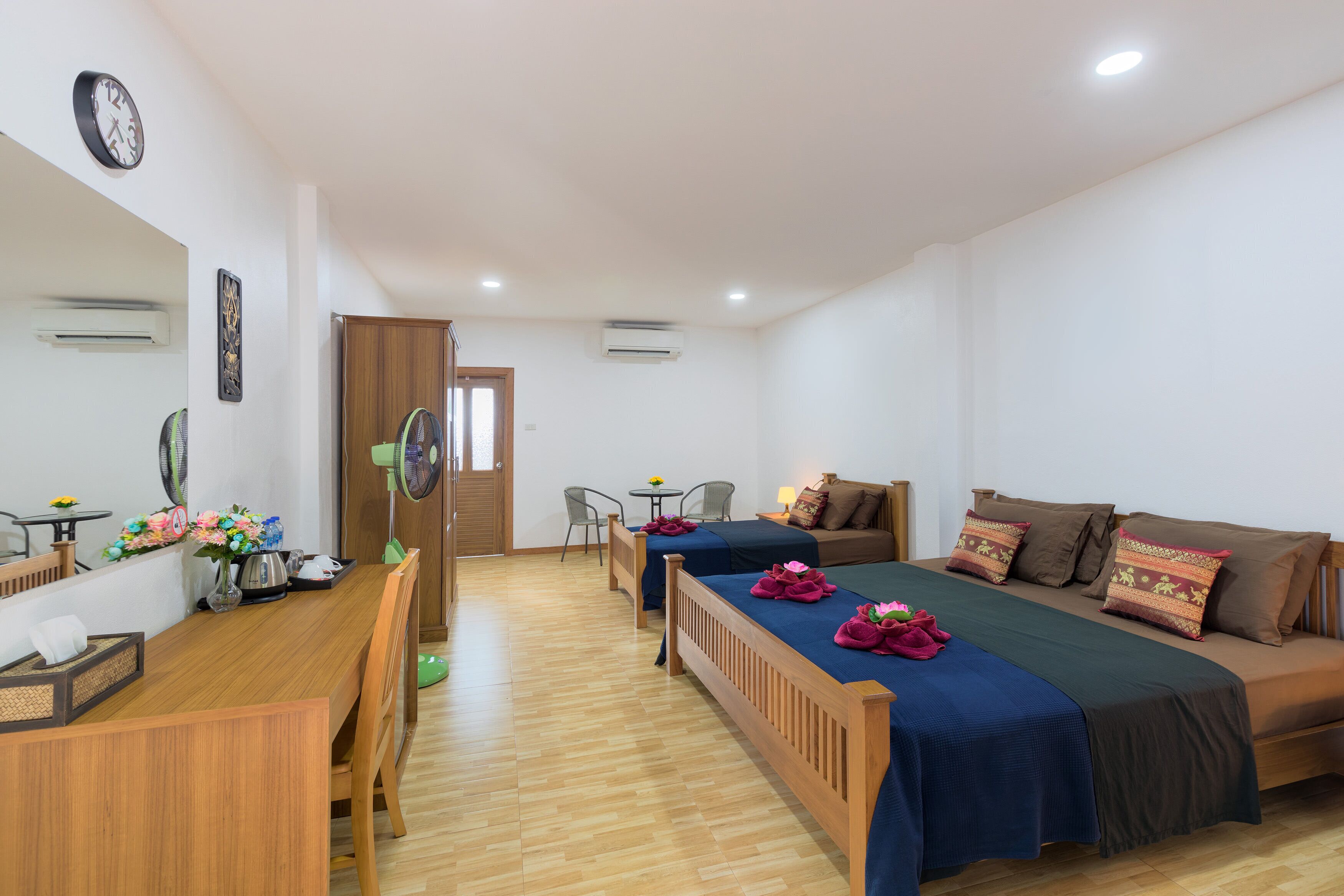 A hotel room with a wooden bed, a wooden desk, a chair, a small table with a lamp, and a clock on the wall.