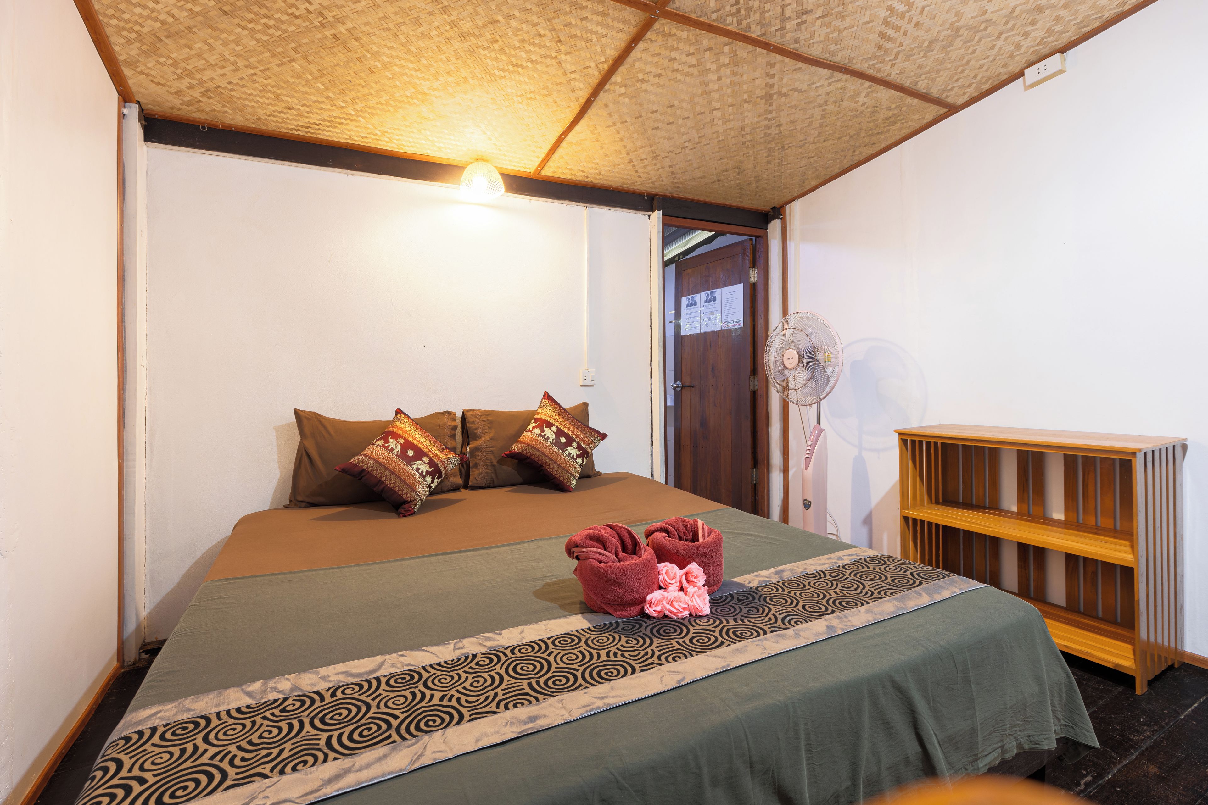 A room with a bed, pillows, a bedside fan, and a wooden shelf.
