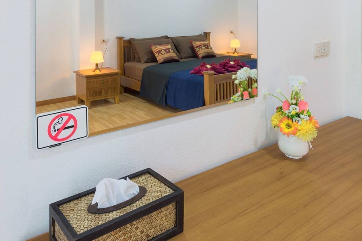 A bedroom with a large mirror, a bed with pillows, a bedside table with a lamp, a no-smoking sign, a small table with a tissue box, and a vase with flowers.
