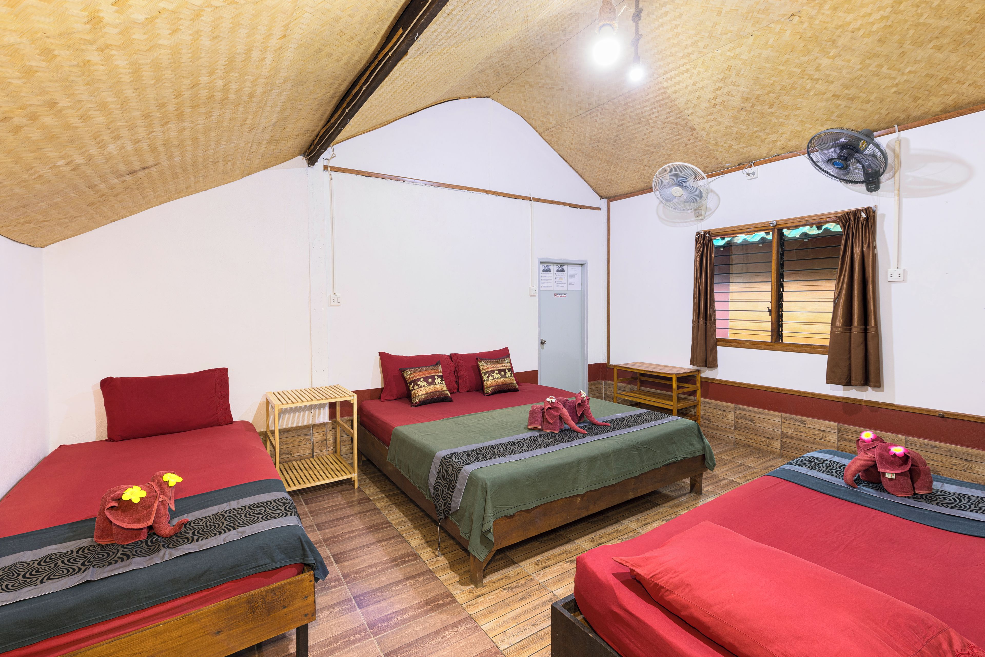 Una habitación con tres camas, piso de madera, una ventana con cortinas y un ventilador de pared.