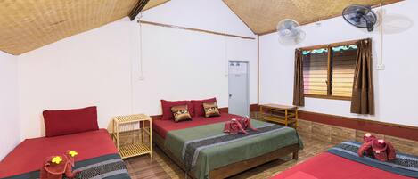 Una habitación con tres camas, piso de madera, una ventana con cortinas y un ventilador de pared.
