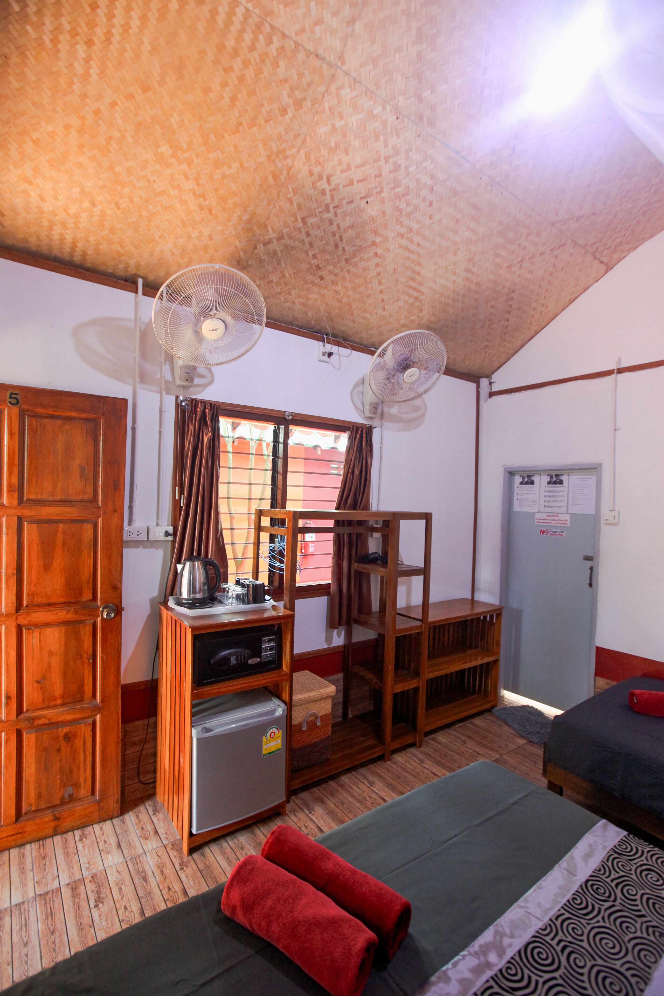 A room with two beds, a wooden wardrobe, a small refrigerator, a desk with a chair, and two ceiling fans.