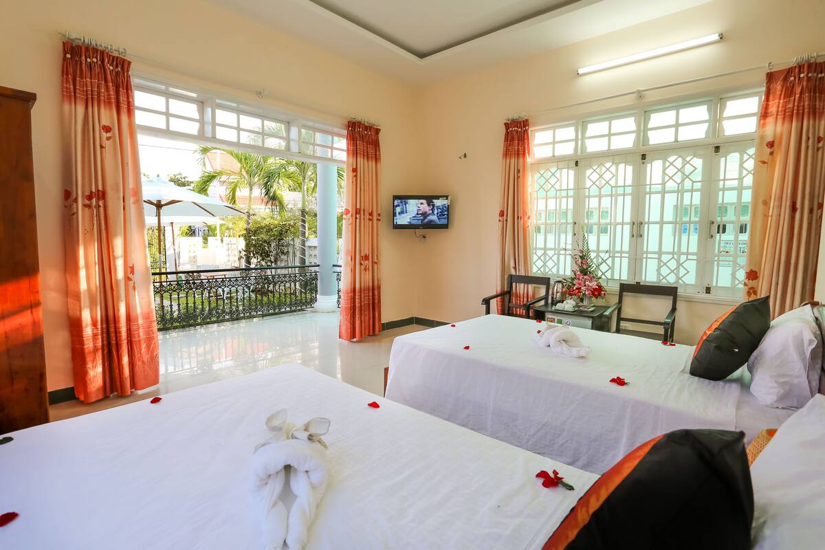 Habitación de hotel con dos camas, televisión y balcón con vista a palmeras.