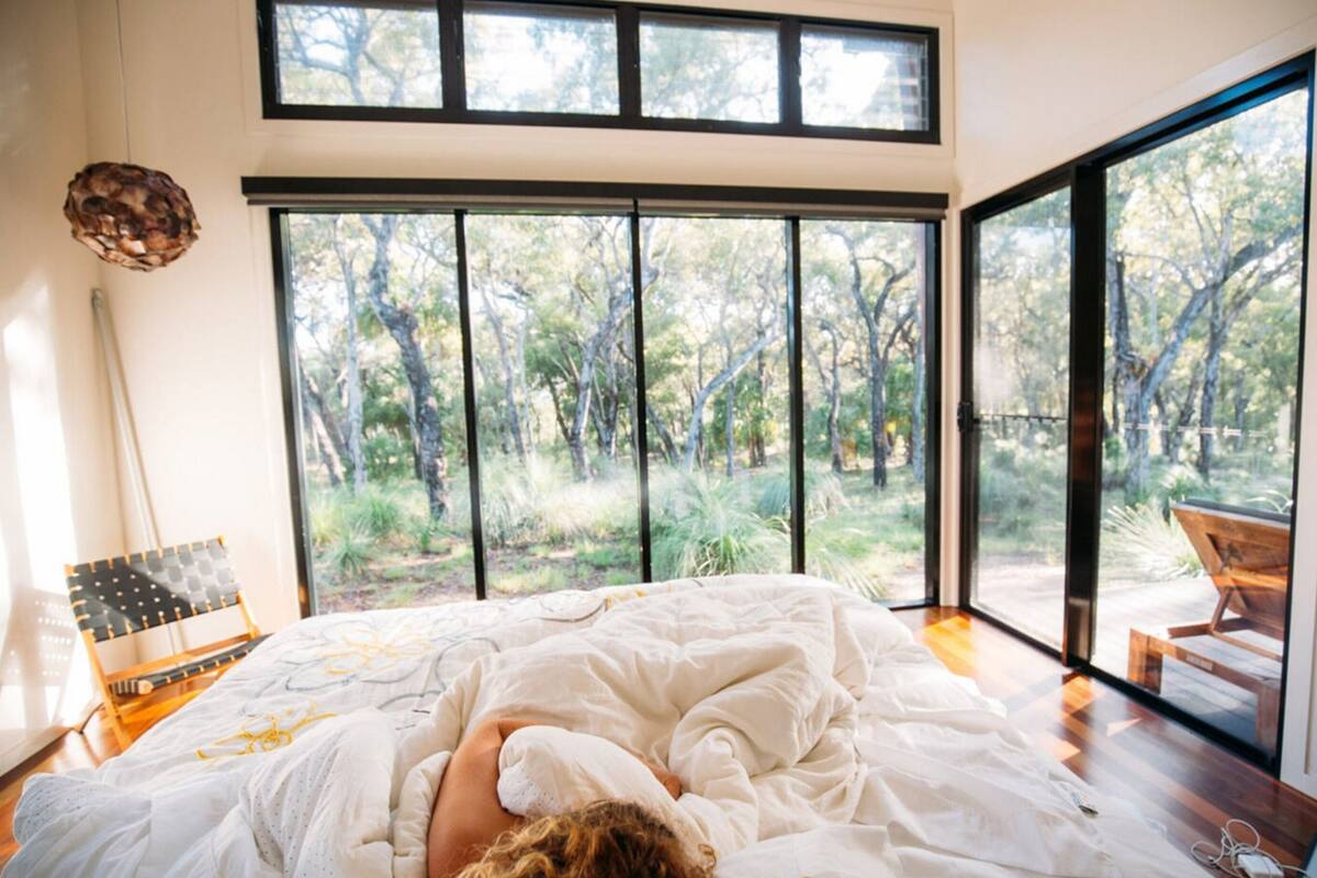 Ein Schlafzimmer mit einem großen Bett, Blick auf einen Garten durch raumhohe Fenster und einer Holzterrasse davor.