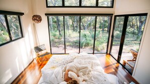 Un dormitorio con un amplio ventanal que da a un bosque, suelo de madera y una cama con edredón blanco.