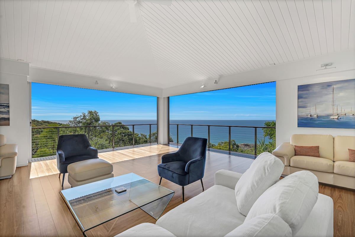 Ein modernes Wohnzimmer mit einem Glastisch, zwei Sesseln und einem Sofa. Große Fenster bieten Blick auf das Meer und die Hügel.