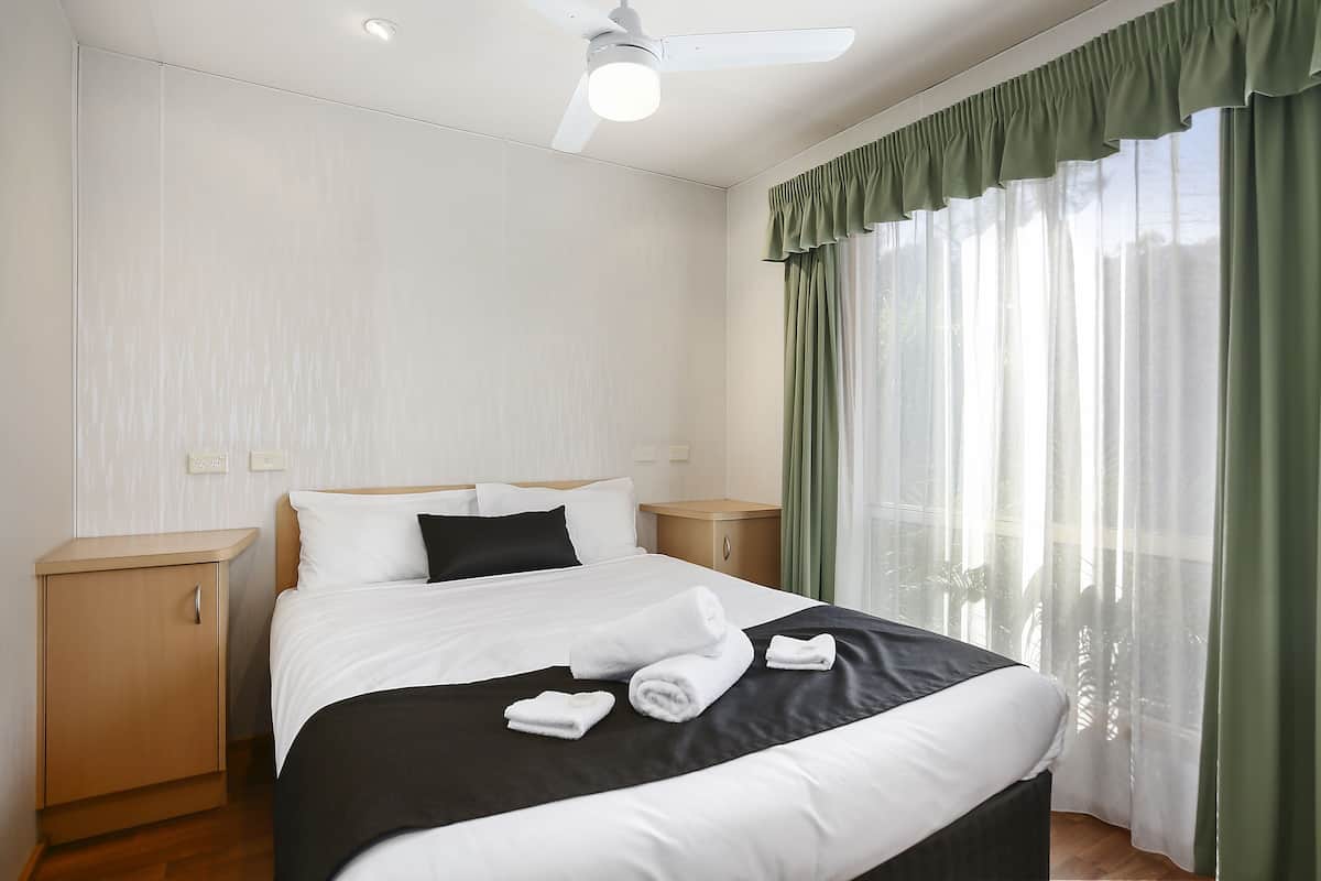 A neatly made bed with white and black bedding, a wooden nightstand, a ceiling fan, and sheer green curtains.
