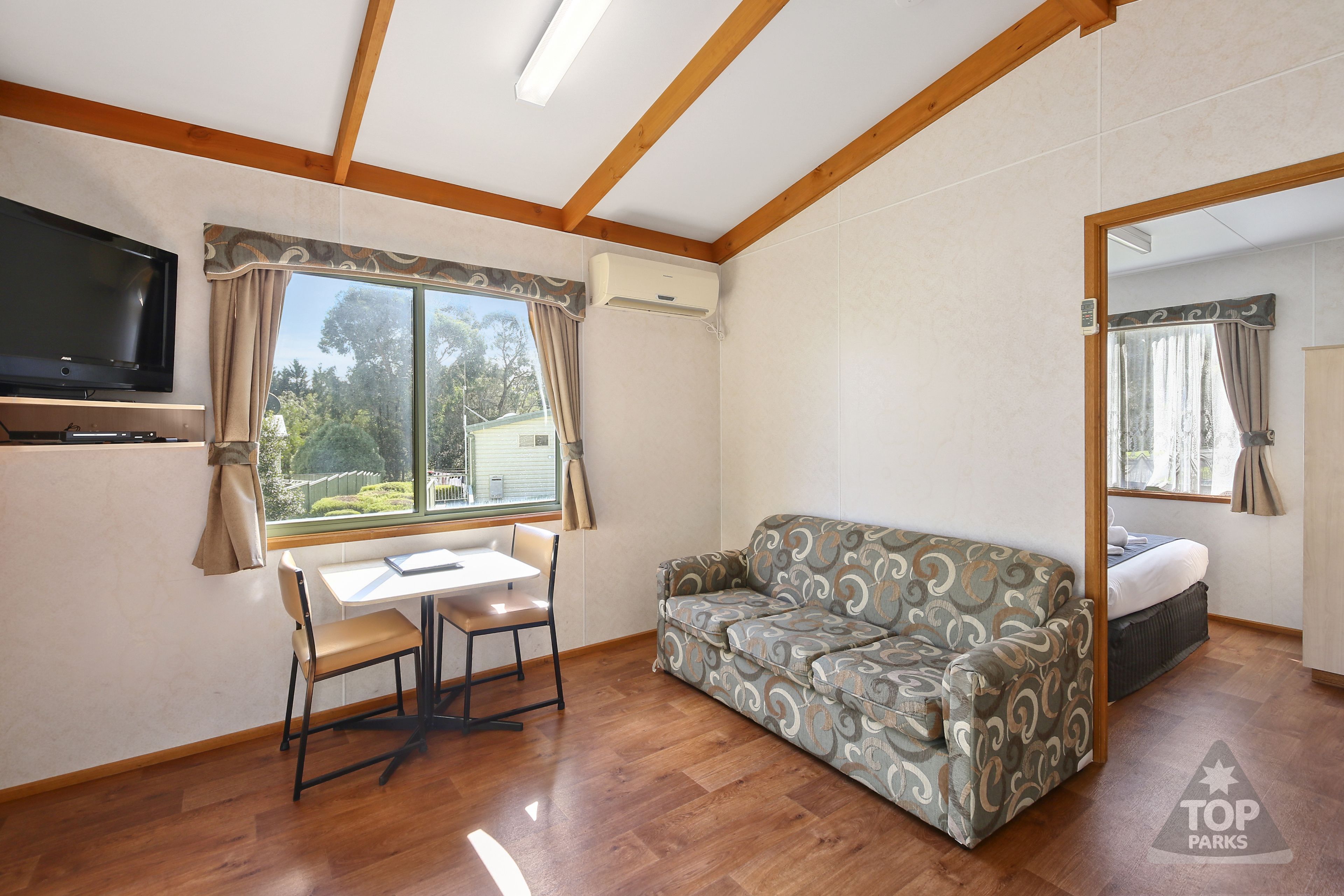 Una habitación con techo de madera, un sofá, una mesa con sillas, un televisor montado en la pared y una ventana con cortinas.