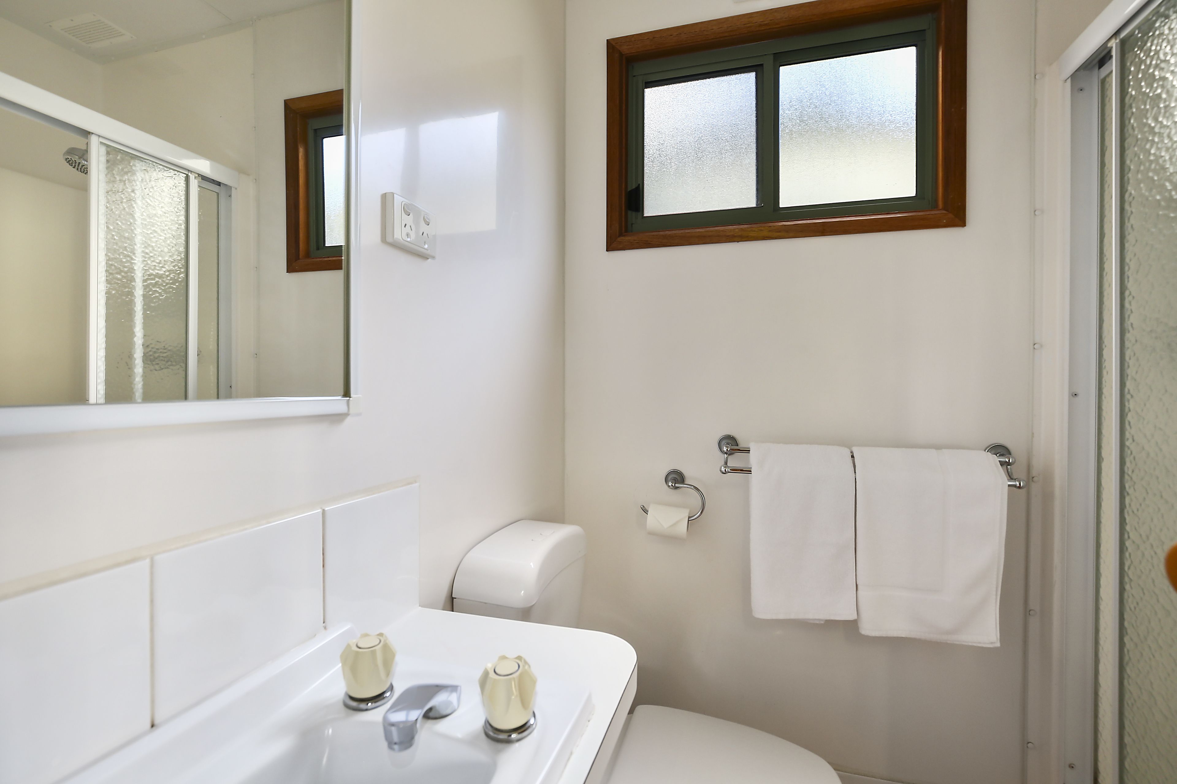 A bathroom with a white sink, a toilet, a mirror, and a window with a wooden frame.