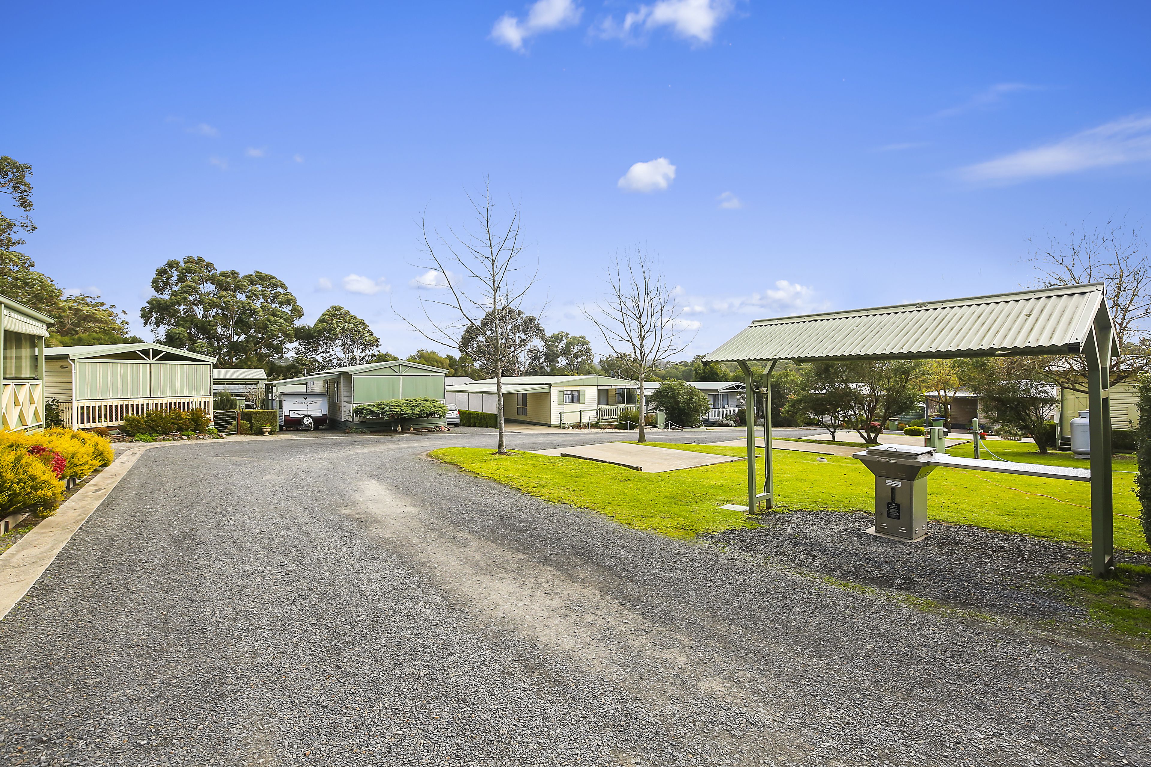 A gravel driveway leading to a row of single-story buildings with a green roof, surrounded by grass and trees.