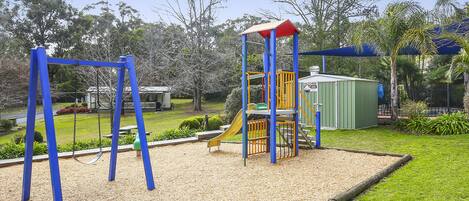 A playground with swings and a slide, surrounded by grass and trees.