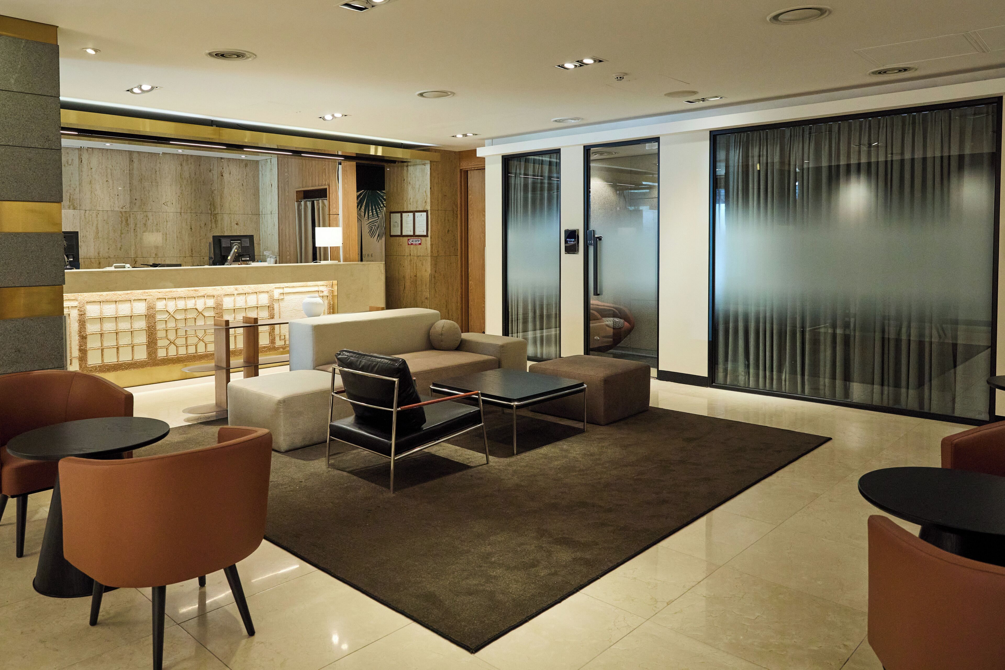 A modern hotel lobby with a reception desk, seating area with sofas and chairs, and a large glass partition.