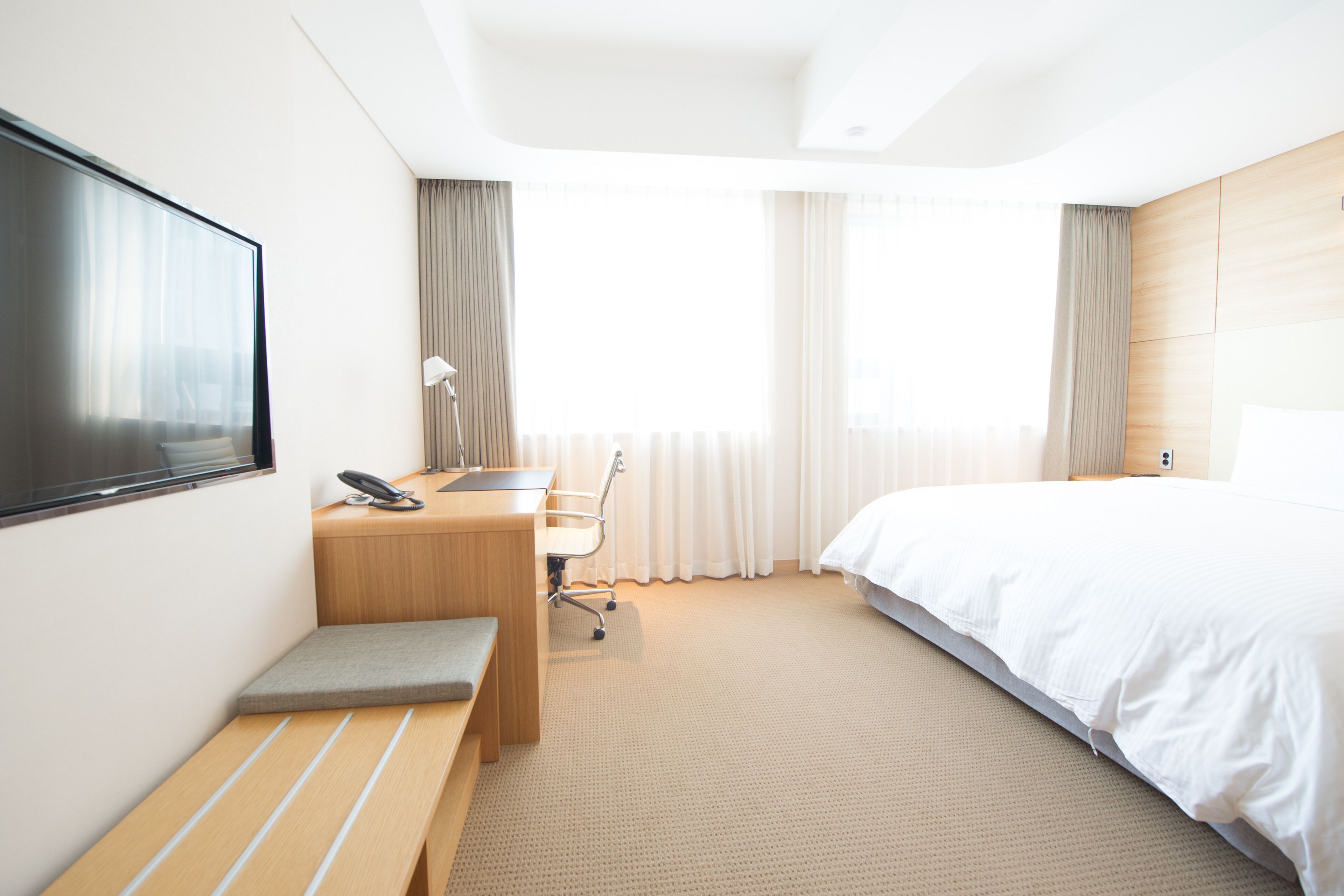 Una habitación de hotel moderna con una cama grande, un escritorio con silla, un televisor montado en la pared y un amplio ventanal con cortinas.